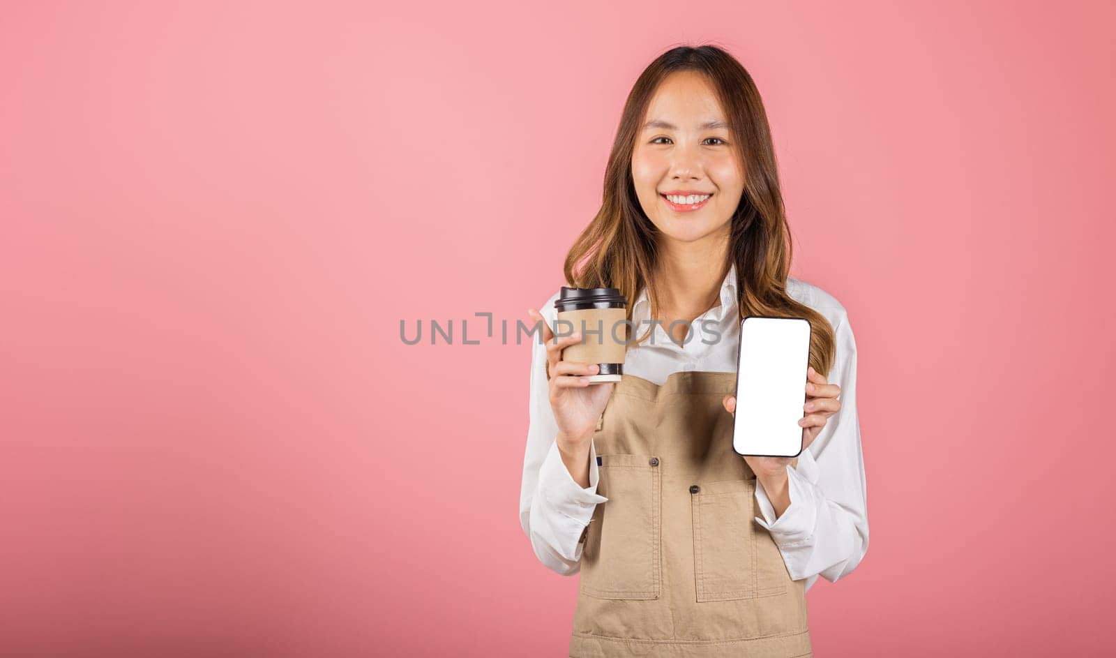 Portrait Asian happy young woman barista bar tender coffee maker holding coffee tea hot cup and mobile phone studio shot isolated on pink background, female smiling hold takeaways cup and smartphone