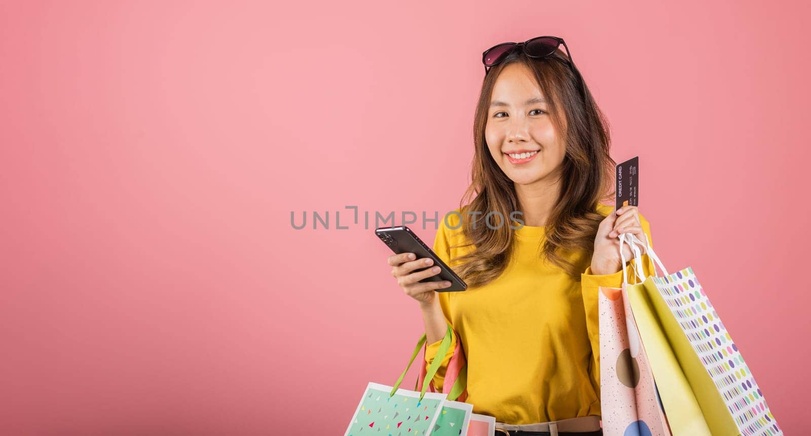 Portrait Asian happy young woman shopper smiling standing excited holding online shopping bags, smartphone and credit card for payment on hand in summer, studio shot isolated by Sorapop