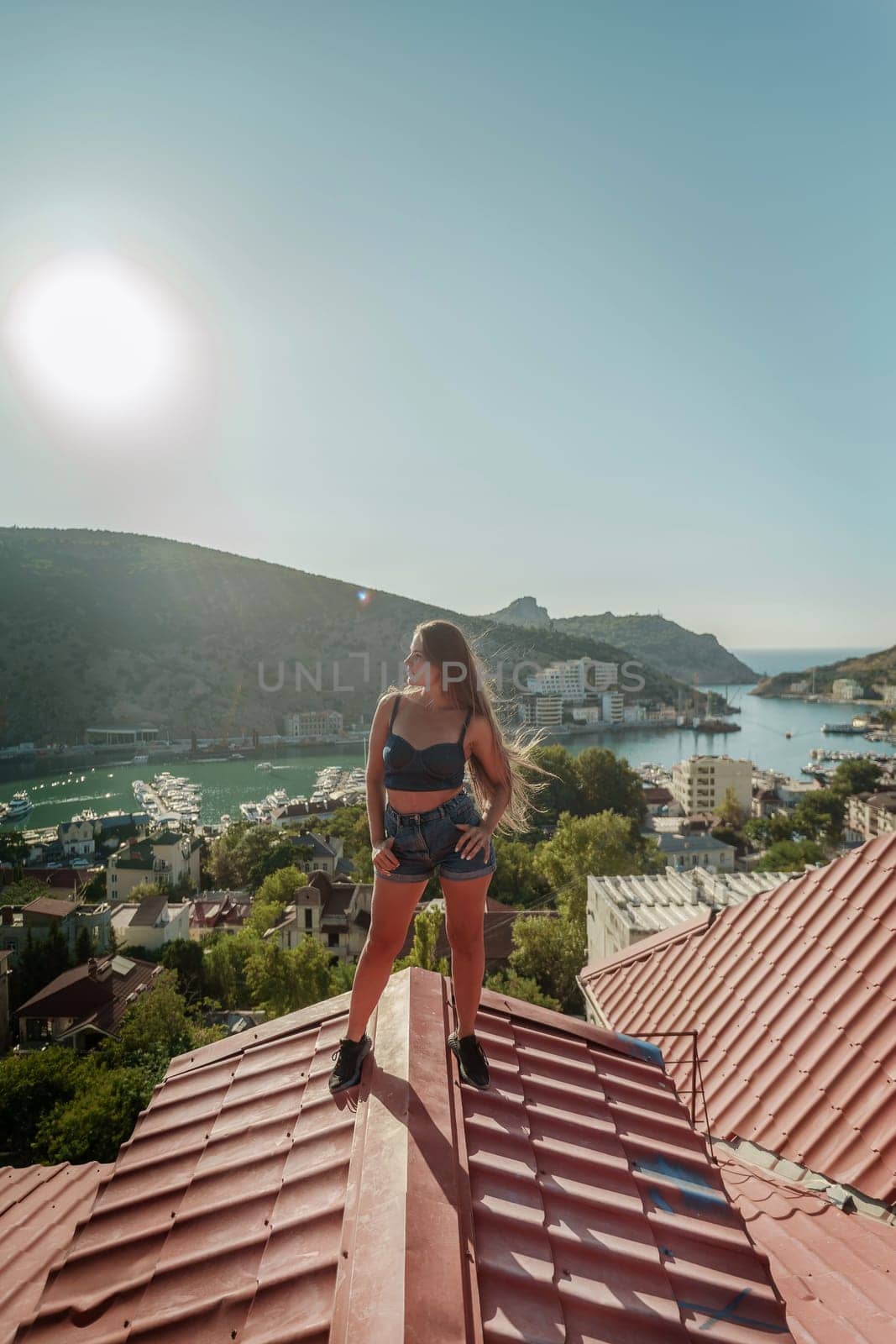 Woman standing on rooftop, enjoys town view and sea mountains. Peaceful rooftop relaxation. Below her, there is a town with several boats visible in the water. Rooftop vantage point