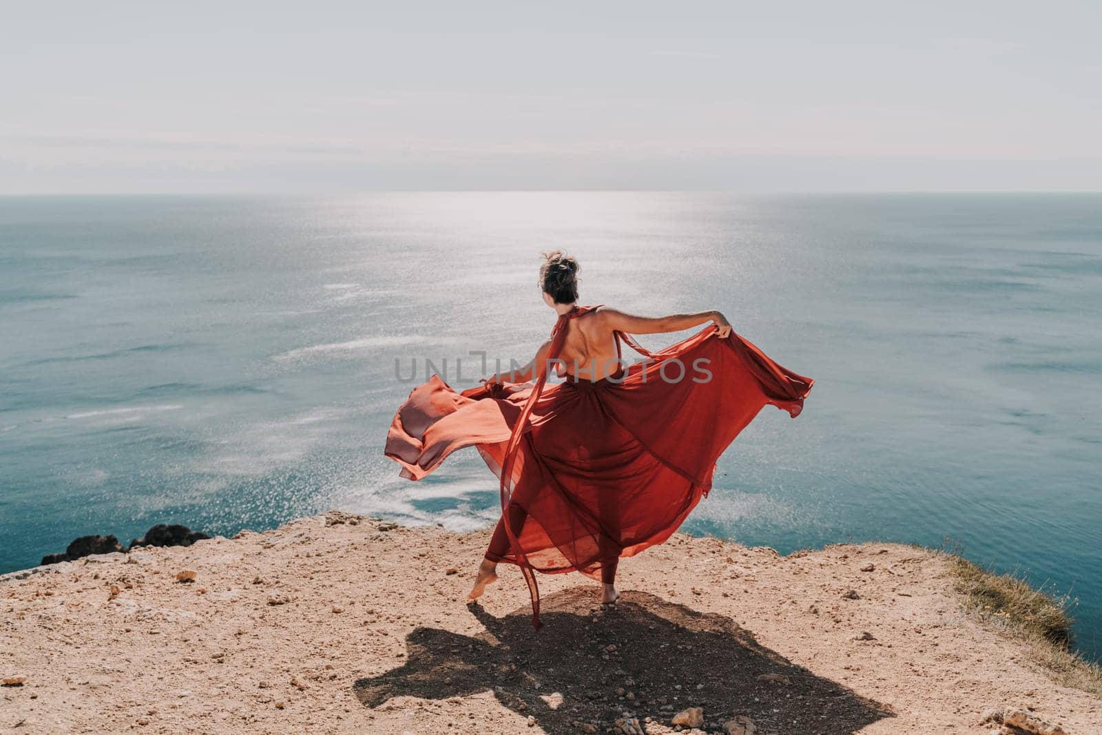 Woman red dress sea. Female dancer posing on a rocky outcrop high above the sea. Girl on the nature on blue sky background. Fashion photo