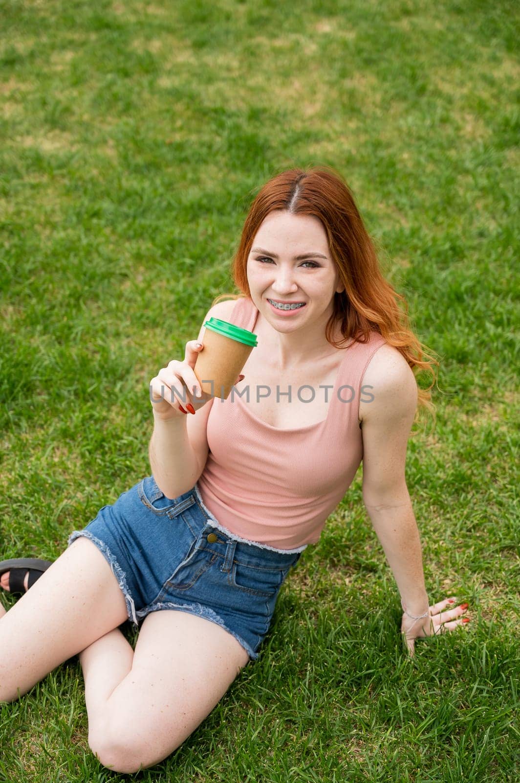 A beautiful young woman with braces on her teeth drinks from a cardboard cup while sitting on the grass. Vertical photo. by mrwed54