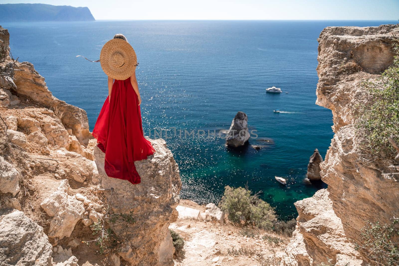 woman in a red dress stands on a cliff overlooking the ocean. The scene is serene and peaceful, with the woman's hat and dress adding a touch of elegance to the image