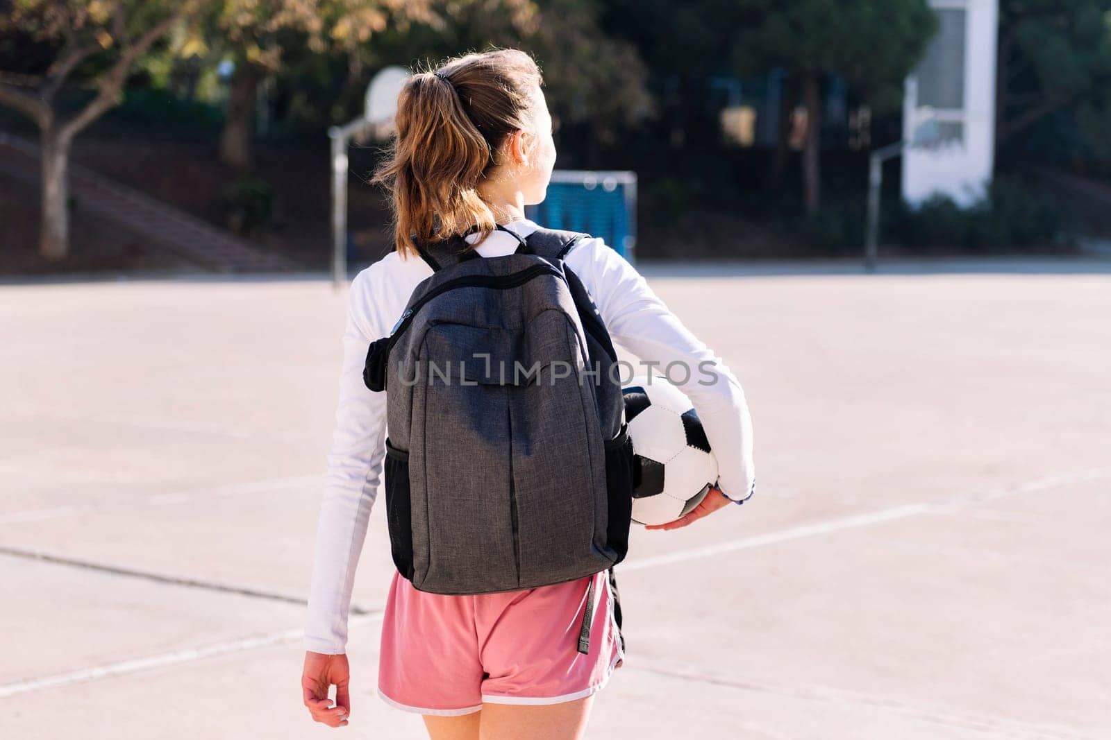 rear view of an unrecognizable young caucasian woman with backpack in a urban football court with a soccer ball in hand, concept of sport and active lifestyle