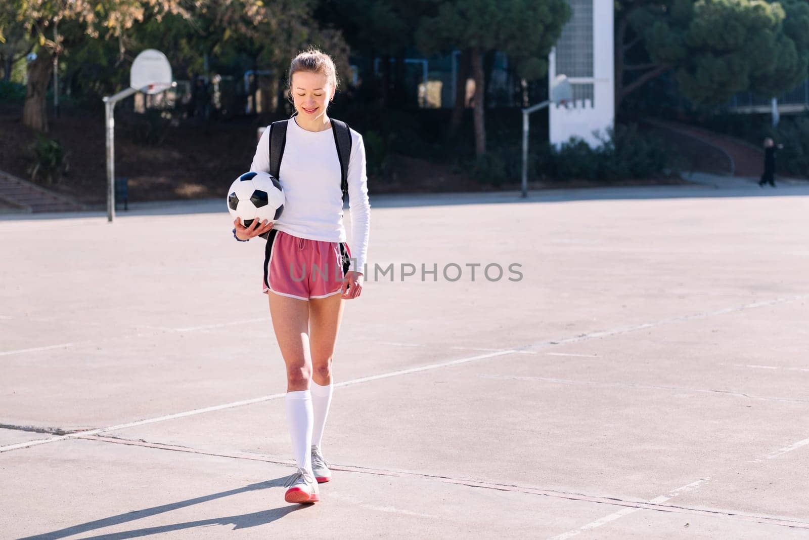 smiling young caucasian woman with backpack walking at urban football court with a soccer ball in hand, concept of sport and active lifestyle, copy space for text