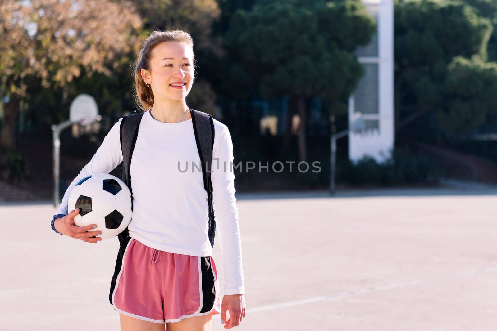 smiling young caucasian woman with backpack walking at urban football court with a soccer ball in hand, concept of sport and active lifestyle, copy space for text