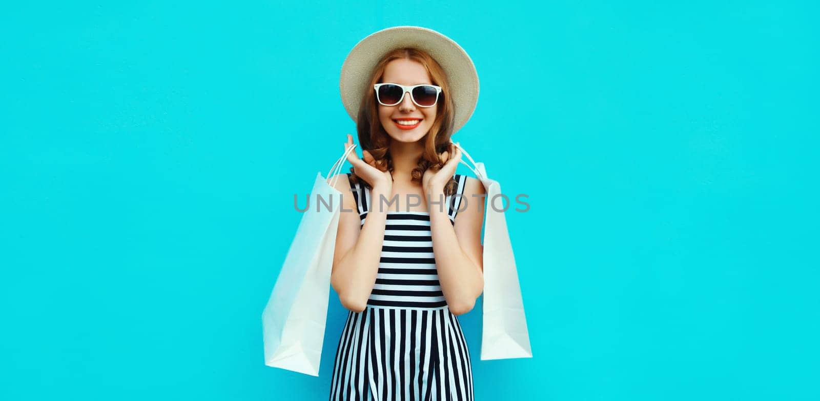 Stylish beautiful happy smiling young woman posing with white shopping bags wearing summer straw hat on blue background