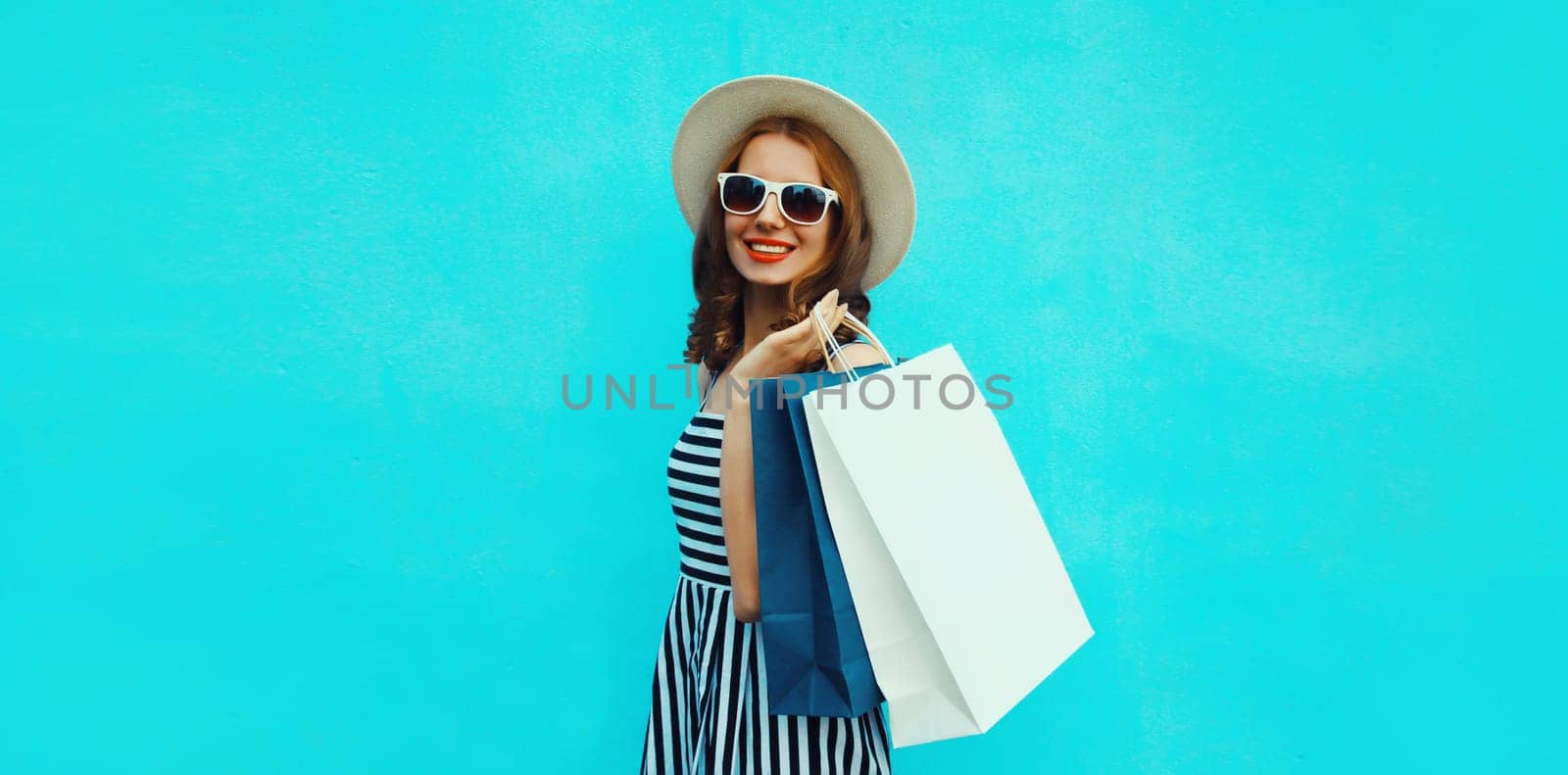 Stylish beautiful happy smiling young woman posing with white shopping bags wearing summer straw hat on blue background