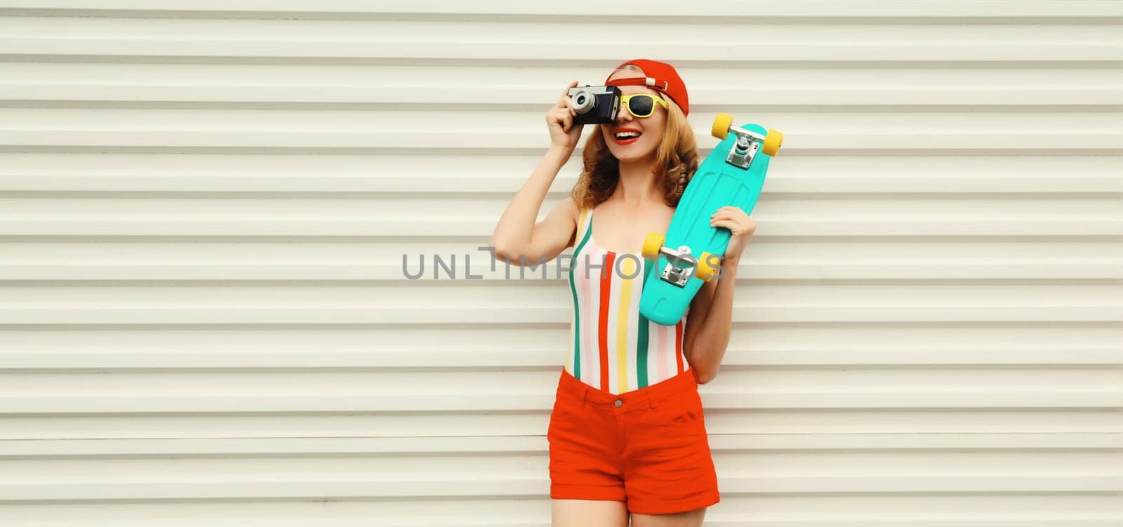 Summer portrait of happy cheerful stylish young woman with skateboard takes picture with camera in colorful clothes on white background