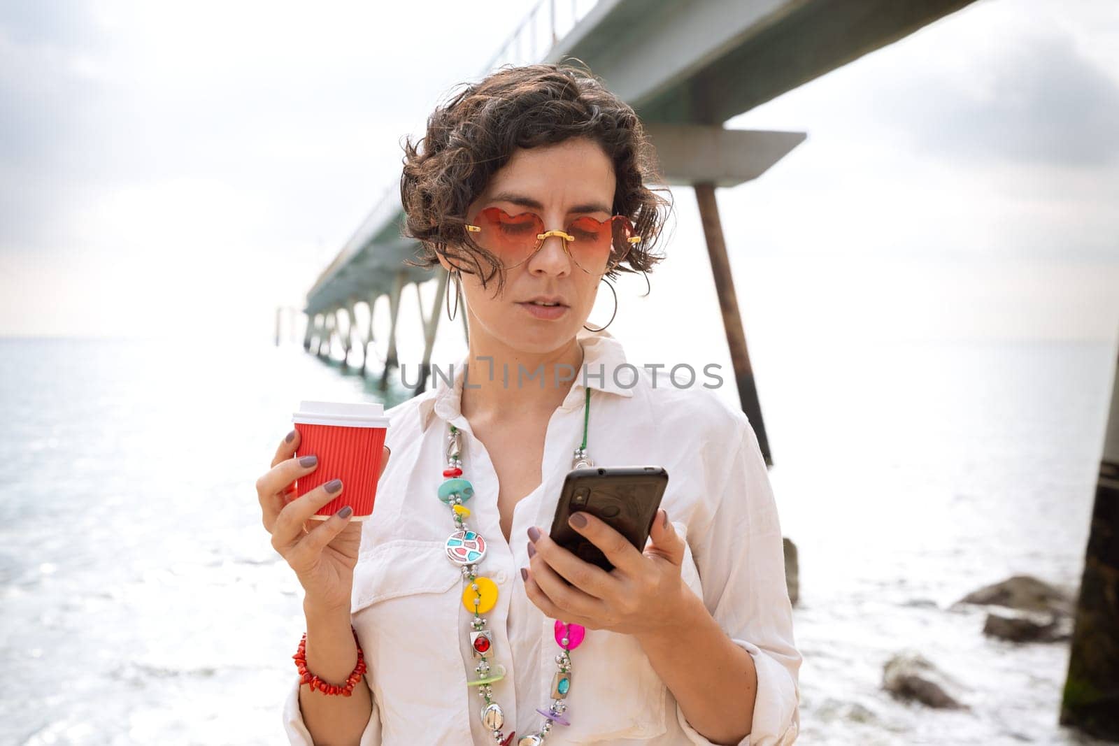 Front view of a caucasian woman wearing sunglasses on vacation by the sea laughing using a social media app with her mobile phone, holding a caffee.