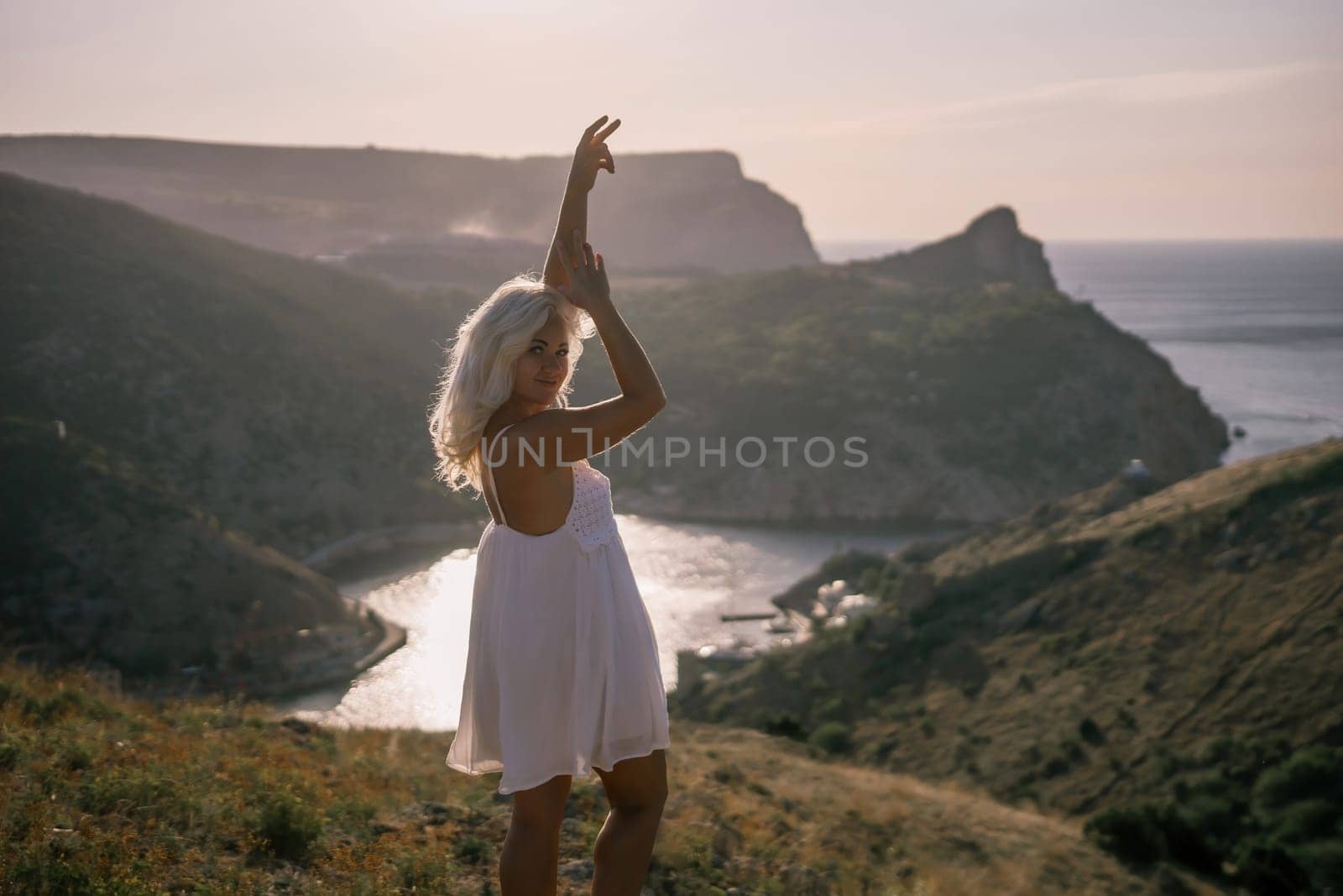 A woman stands on a hill overlooking a body of water. She is wearing a white dress and she is enjoying the view