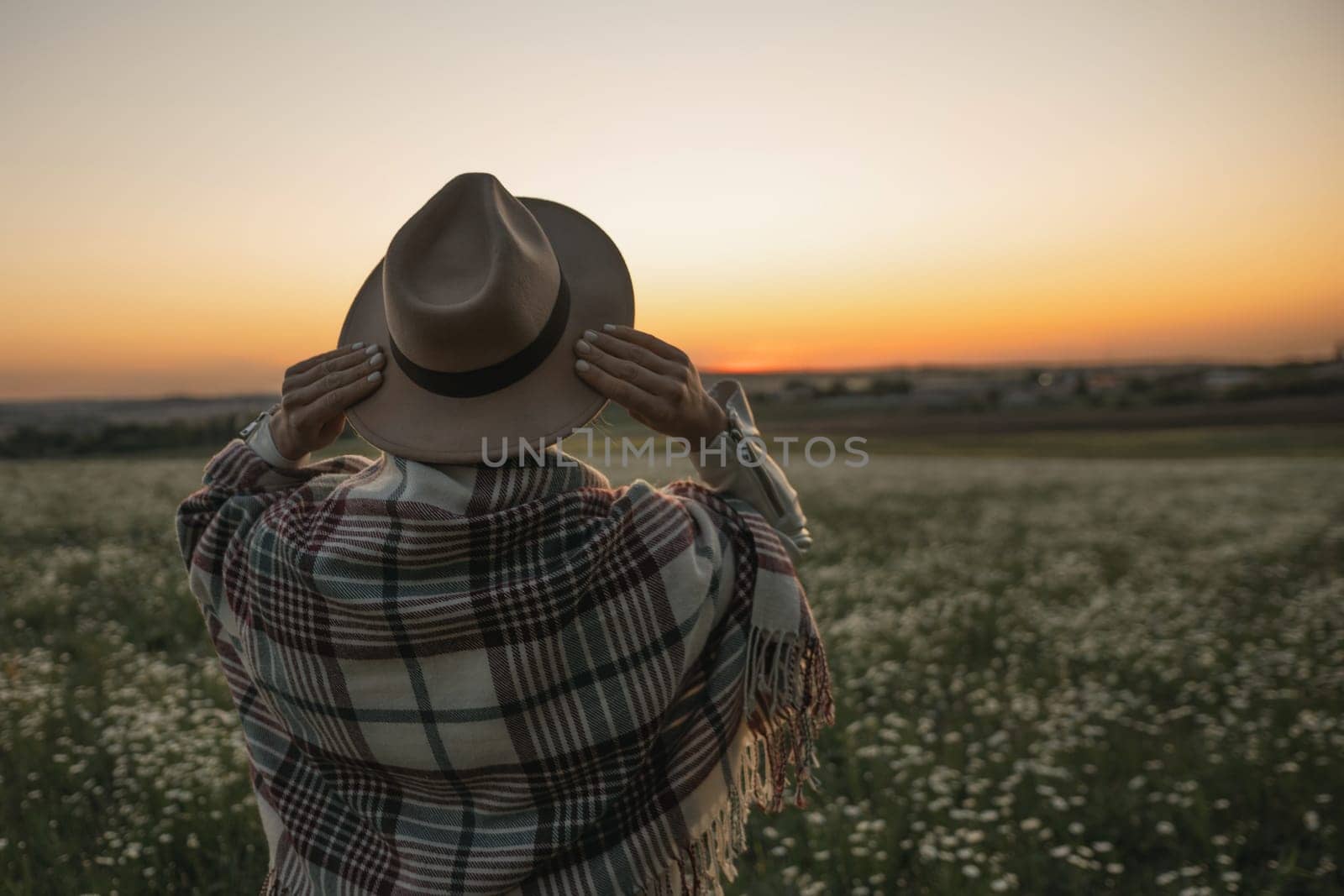A woman wearing a hat and a plaid blanket stands in a field of flowers. The sky is orange and the sun is setting