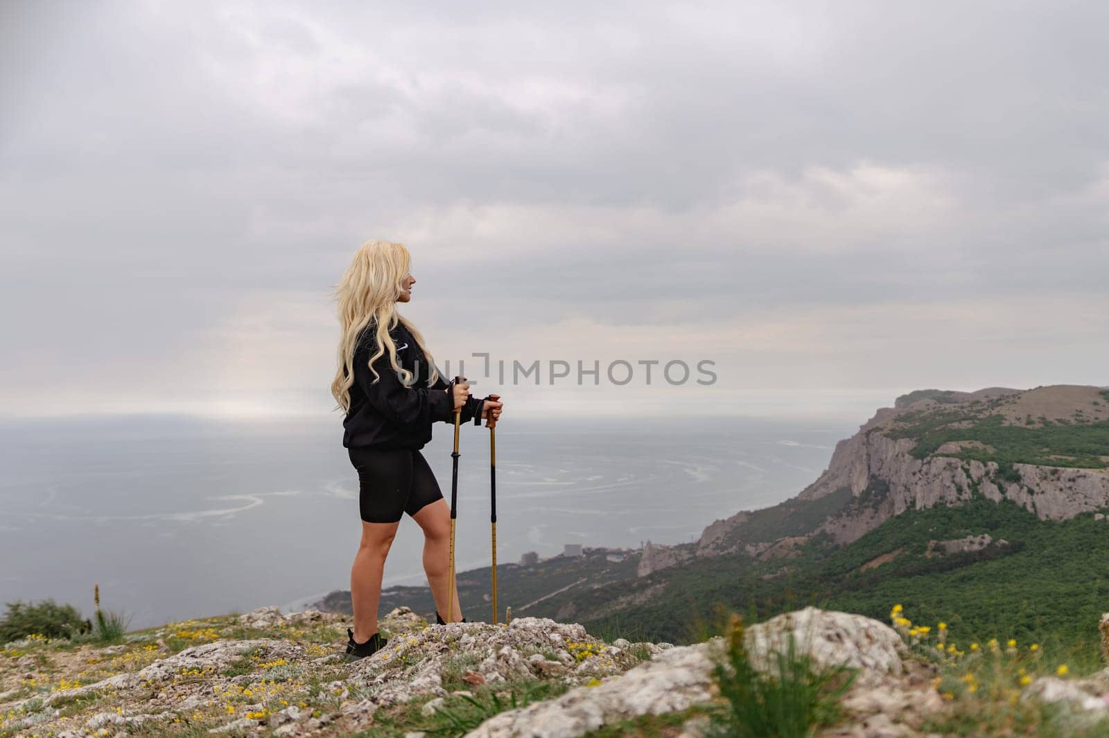 A woman stands on a hill overlooking the ocean. She is wearing a black jacket and shorts. The sky is cloudy and the ocean is in the background