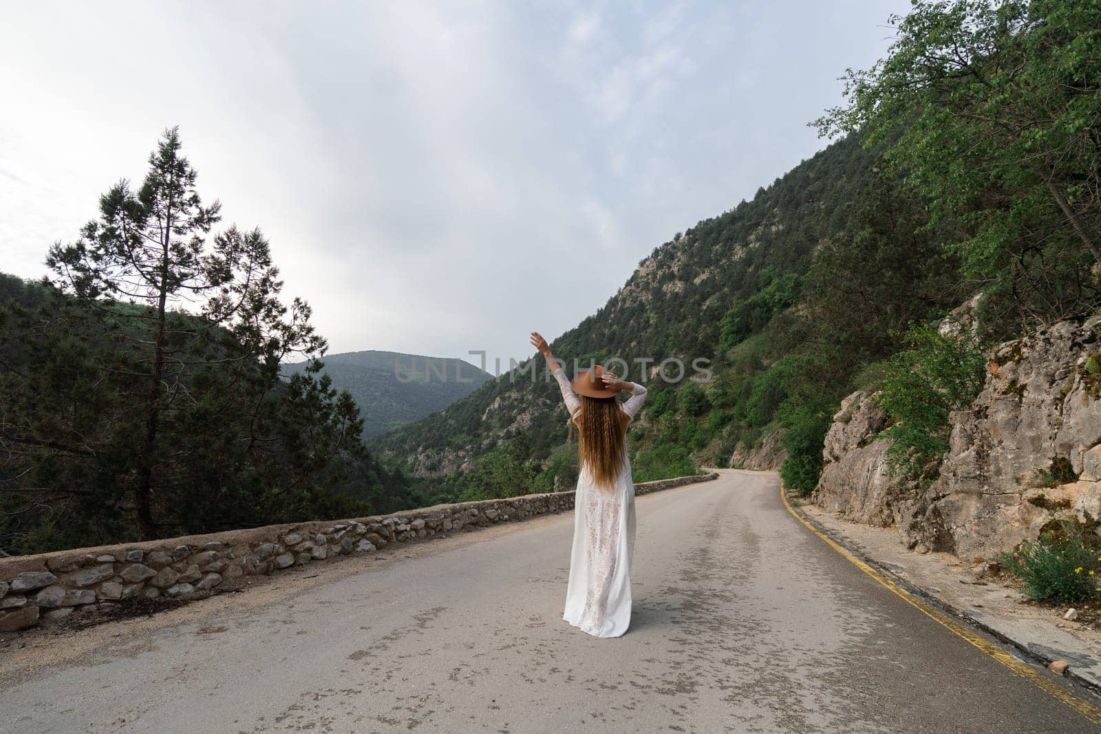 A woman in a white dress is standing on a road, waving her hand. The scene is set in a mountainous area, with trees in the background. The woman is enjoying the view and the fresh air