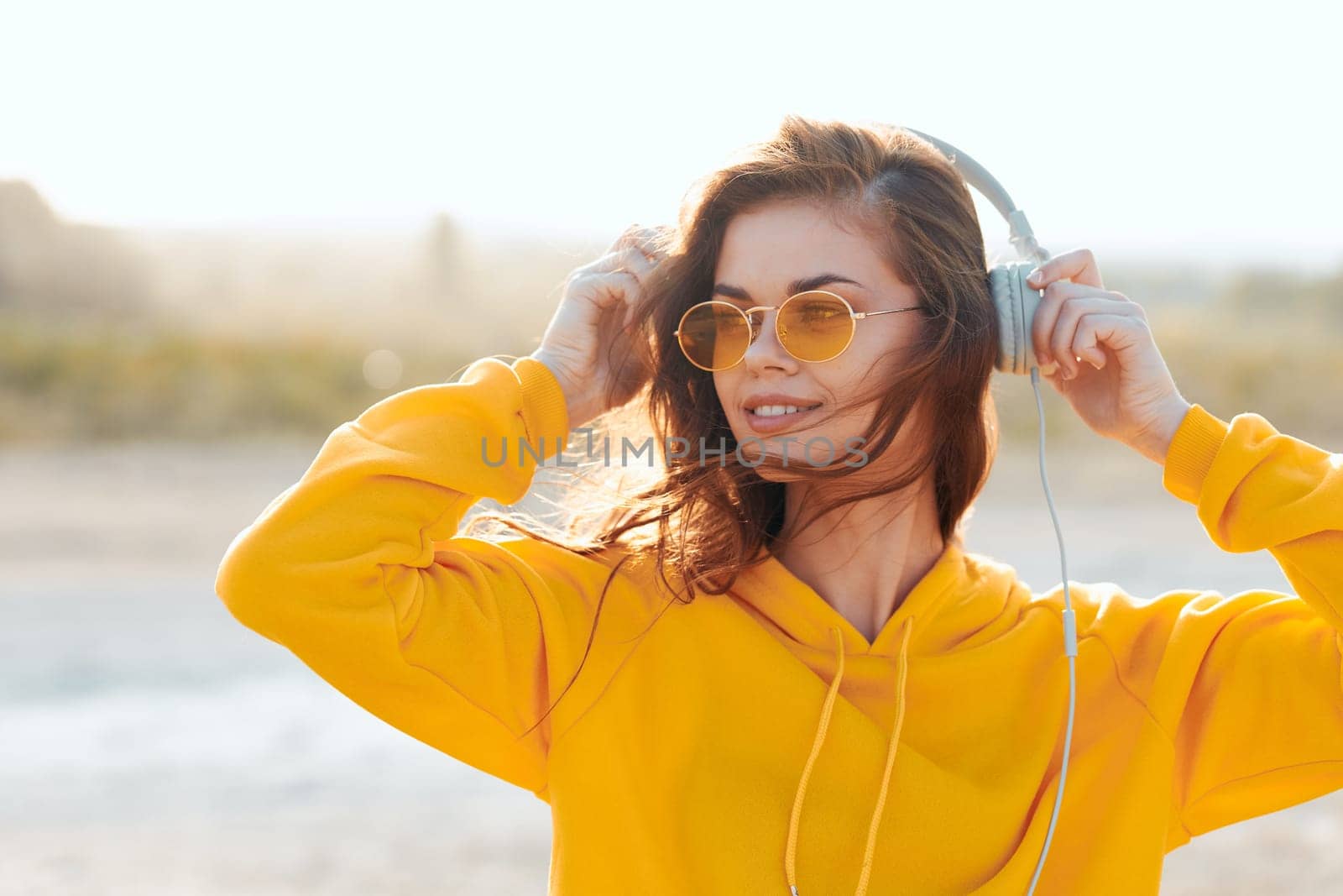 Stylish woman in yellow sweater and sunglasses enjoying music with headphones under the sun by Vichizh