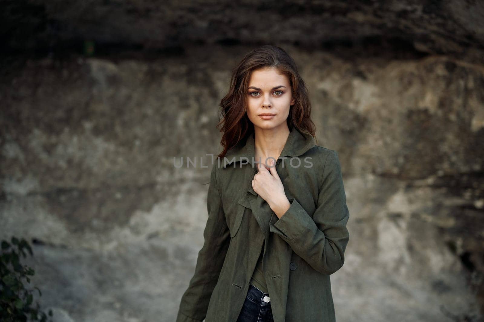 Confident woman in green jacket and jeans standing in front of rugged rock wall with hands on hips