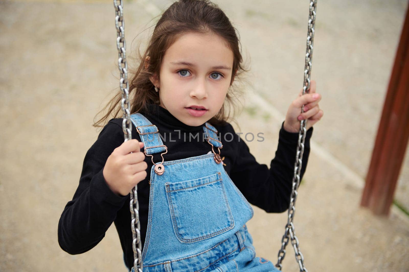 Close-up cute little child girl in denim sundress, looking confidently at camera while swinging on the outdoors playground. People. Happy carefree childhood concept. Lifestyle.