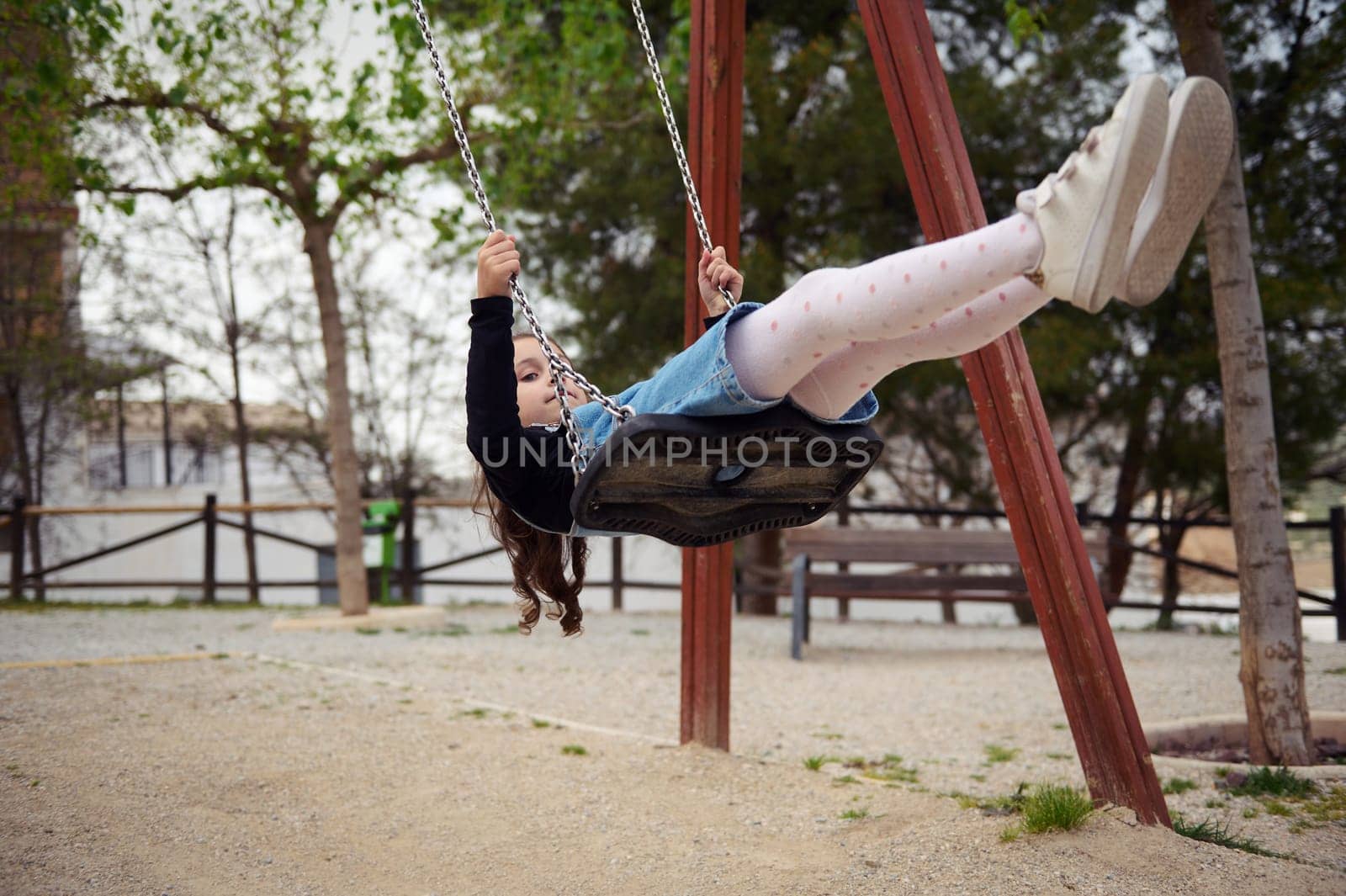 Caucasian cute child girl in casual denim, smiling looking away, relaxing while swinging on the outdoors playground. People. Happy carefree childhood concept. Lifestyle