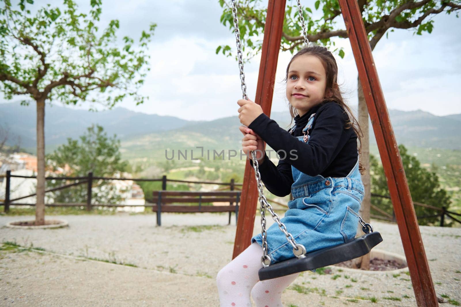 Little child girl 6 years old smiles at camera while sitting on a wooden swing in the playground outdoors. People. Happy carefree childhood. Recreation