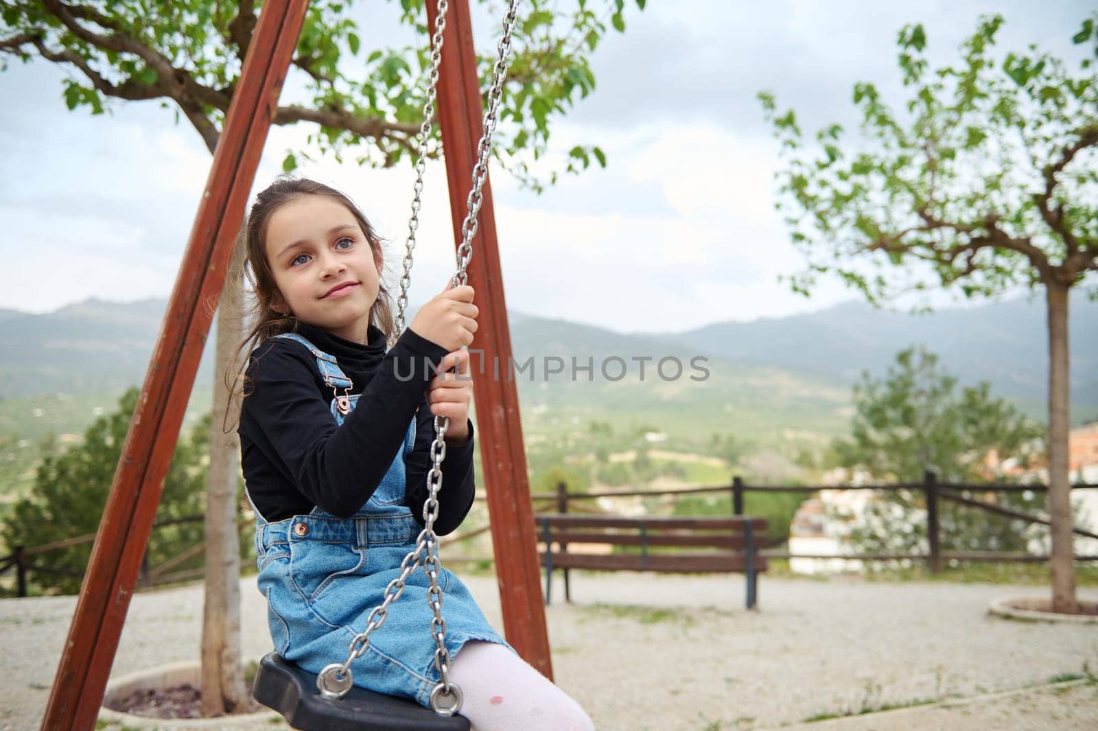 Little child girl smiling and dreamily looking away while sitting on a wooden swing in the playground outdoors. People. Happy carefree childhood. Recreation. Kids development and entertainment