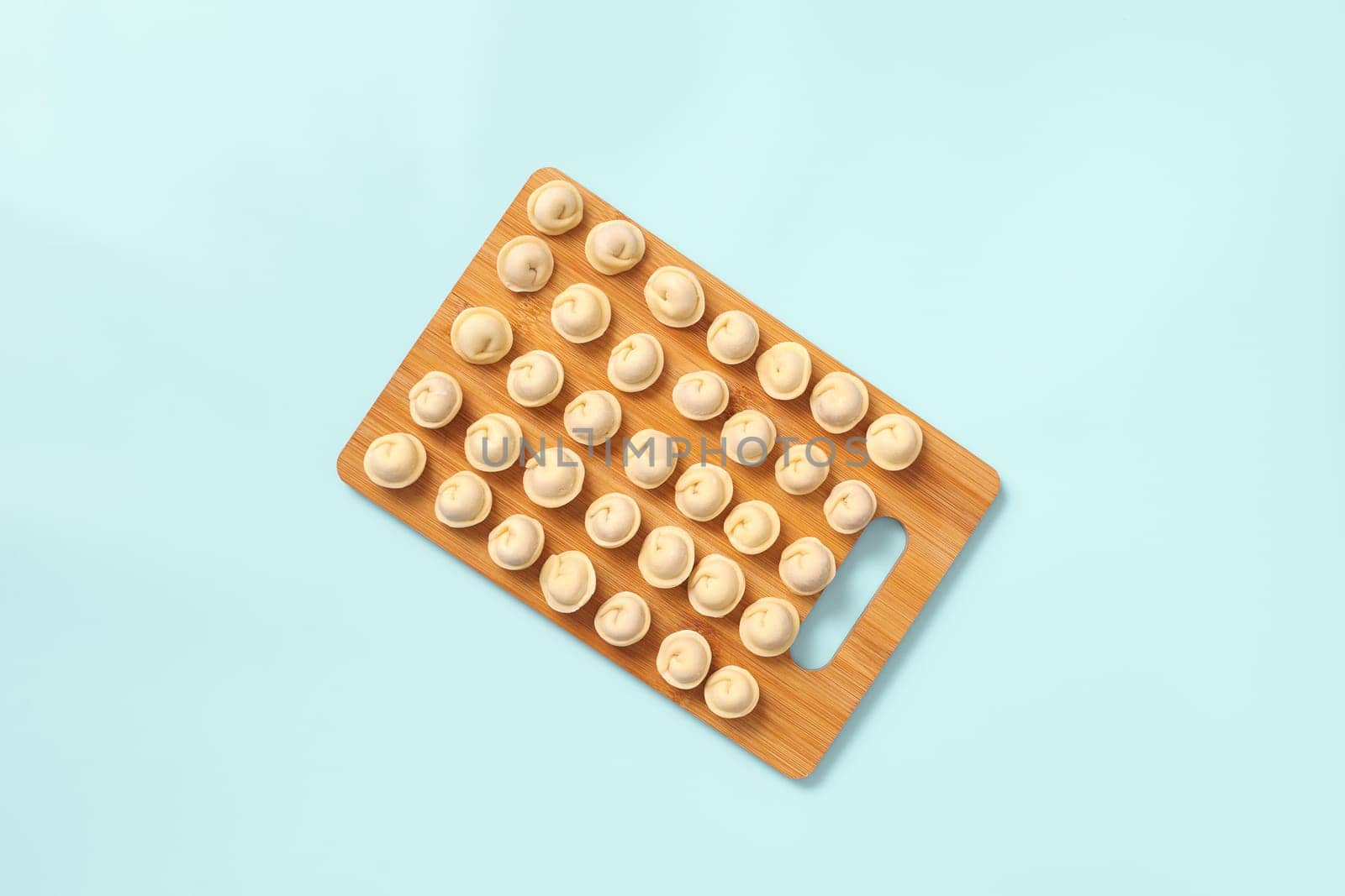 Top view of a wooden board with neatly laid out semi-cooked dumplings on blue background.