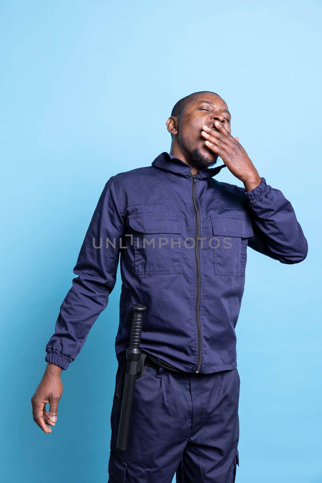 African american security agent feels tired after a long night shift, working on safeguarding a property. Safety officer feeling exhausted and yawning against blue background.