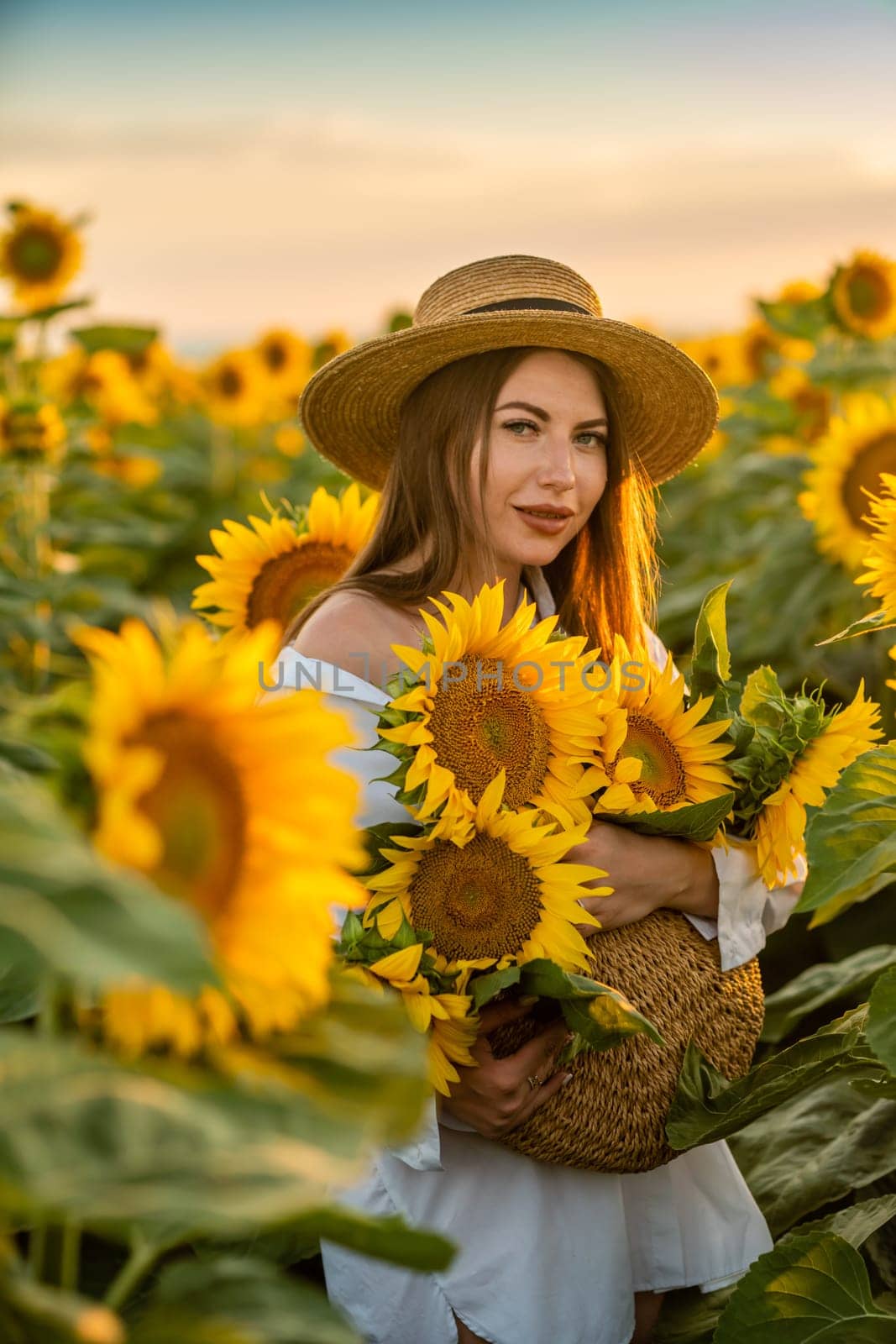 A girl in a hat on a beautiful field of sunflowers against the sky in the evening light of a summer sunset. Sunbeams through the flower field. Natural background