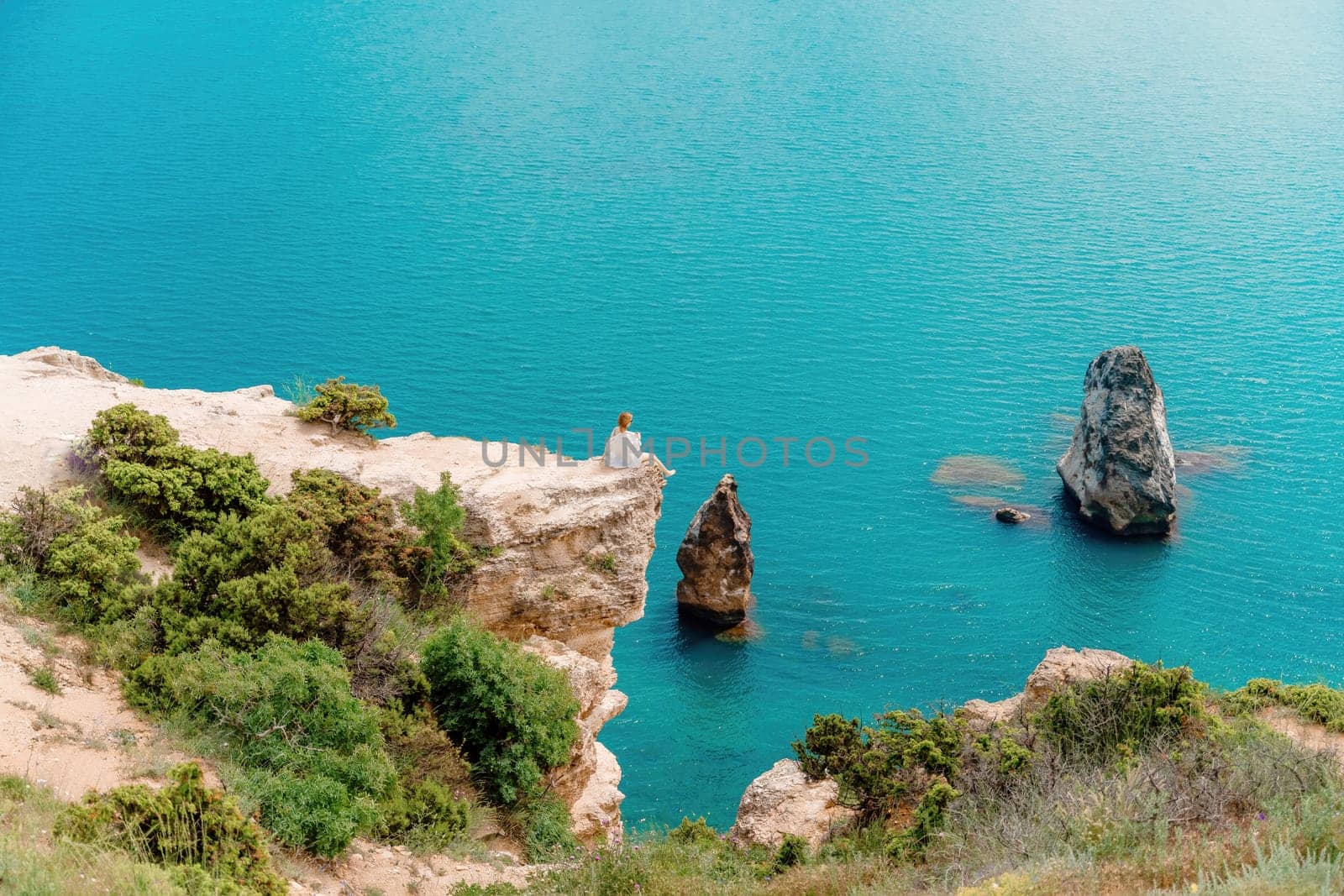 A woman sits on a rock overlooking the ocean. The water is blue and calm. The scene is peaceful and serene