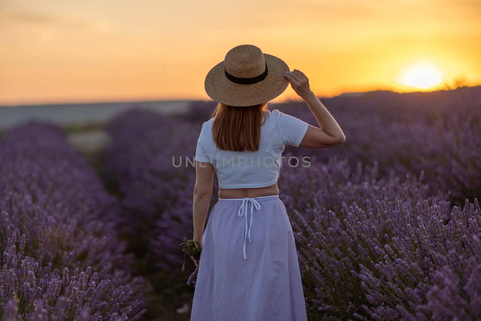 A woman wearing a straw hat stands in a field of lavender. The sun is setting in the background, casting a warm glow over the scene. The woman is enjoying the peaceful atmosphere