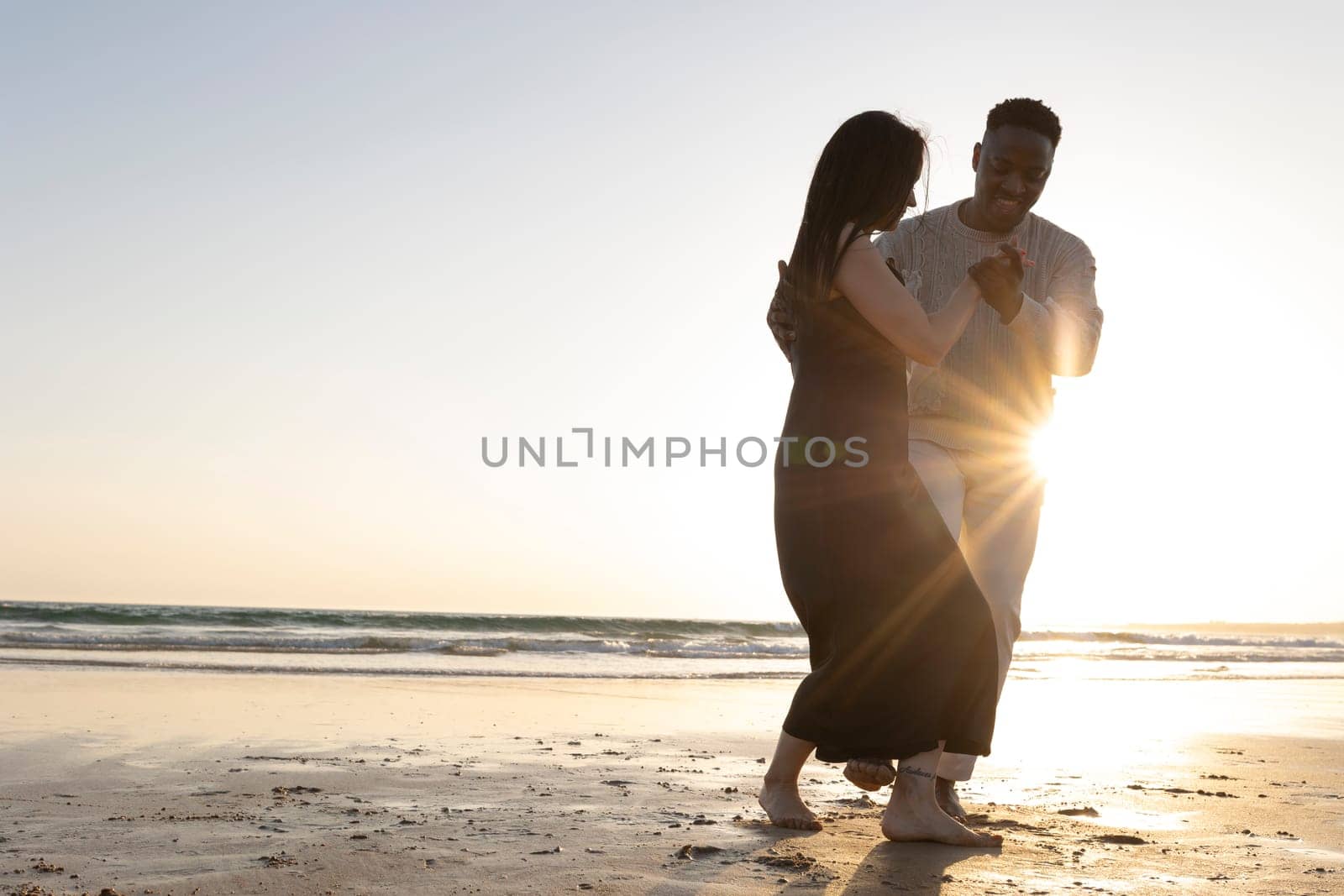 A couple dancing on the beach in the sun. The man is wearing a white shirt and the woman is wearing a black dress