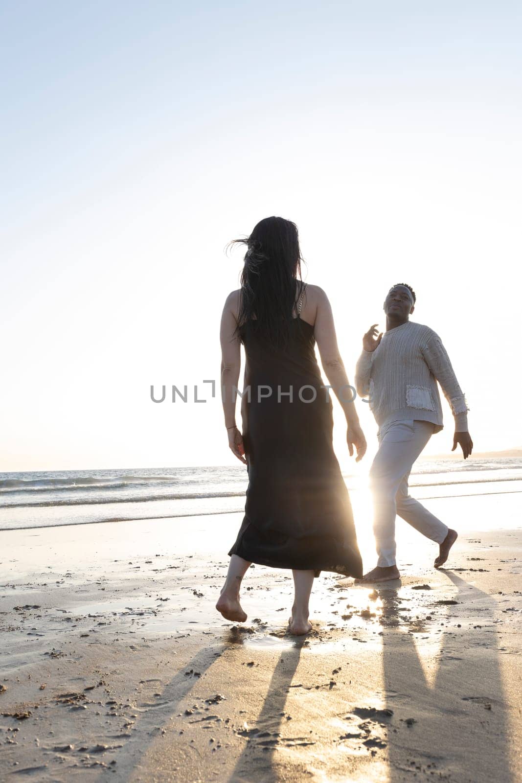 A woman and a man are walking on the beach. The woman is wearing a black dress and the man is wearing a white sweater. The sun is setting in the background, creating a warm and peaceful atmosphere