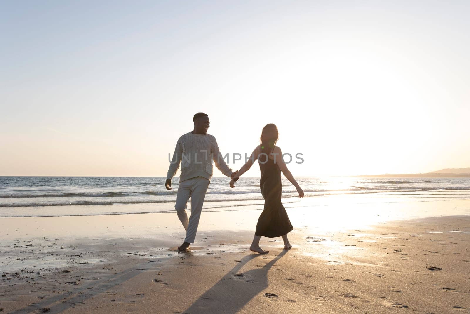 A couple is walking on the beach holding hands. The woman is wearing a black dress. The sun is setting in the background