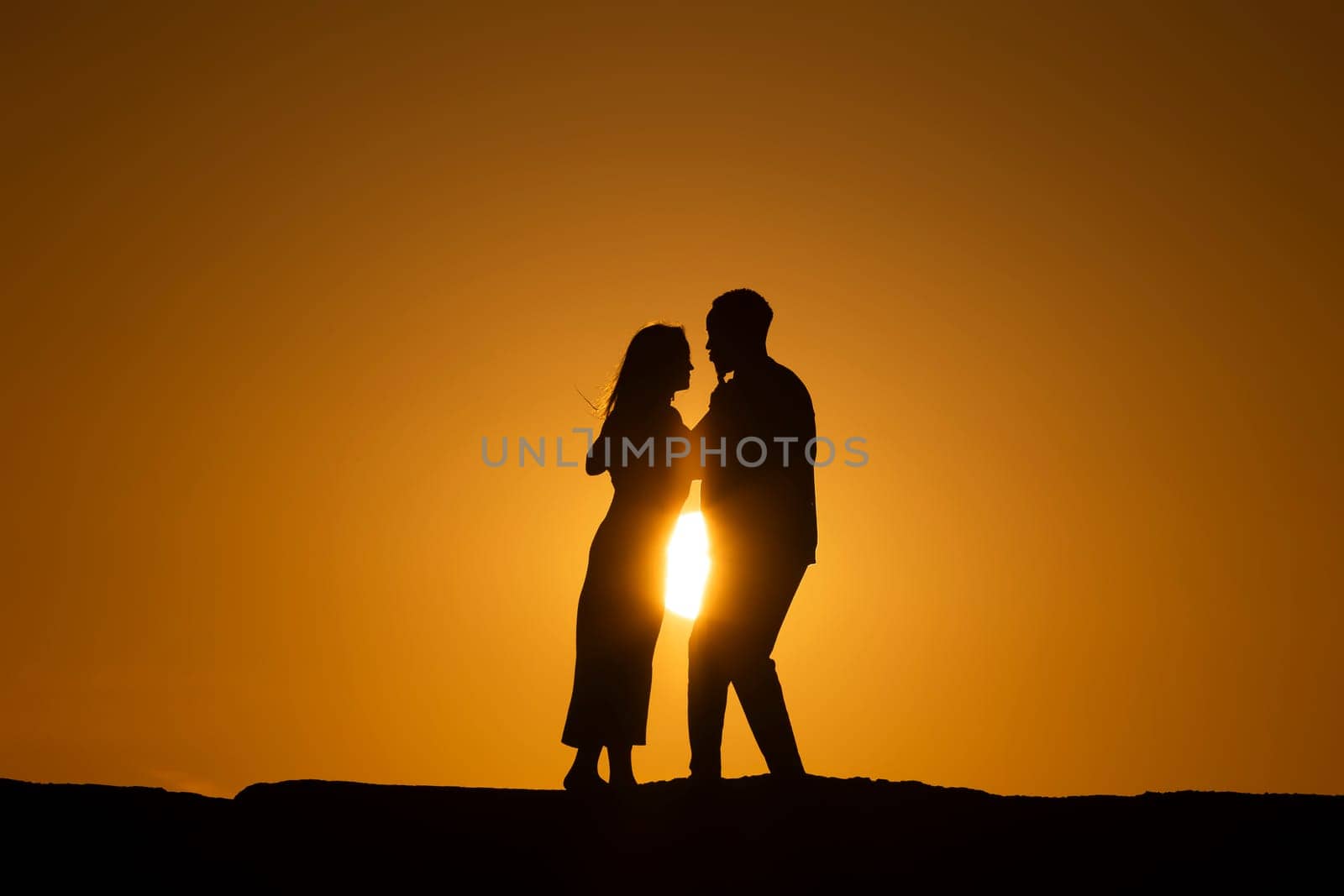 A couple is silhouetted against a sunset, with the woman wearing a dress and the man wearing a jacket
