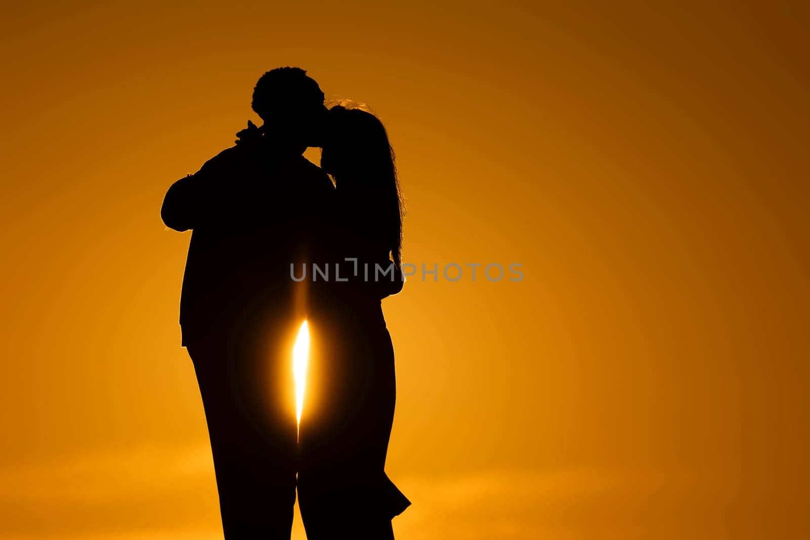 A couple kissing in the sunset. The man is kissing the woman on the cheek. The sun is shining on them, creating a warm and romantic atmosphere