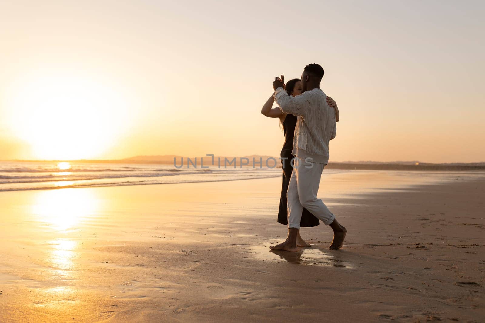A couple is dancing on the beach at sunset. The man is holding the woman's hand and they are both smiling. Scene is romantic and happy
