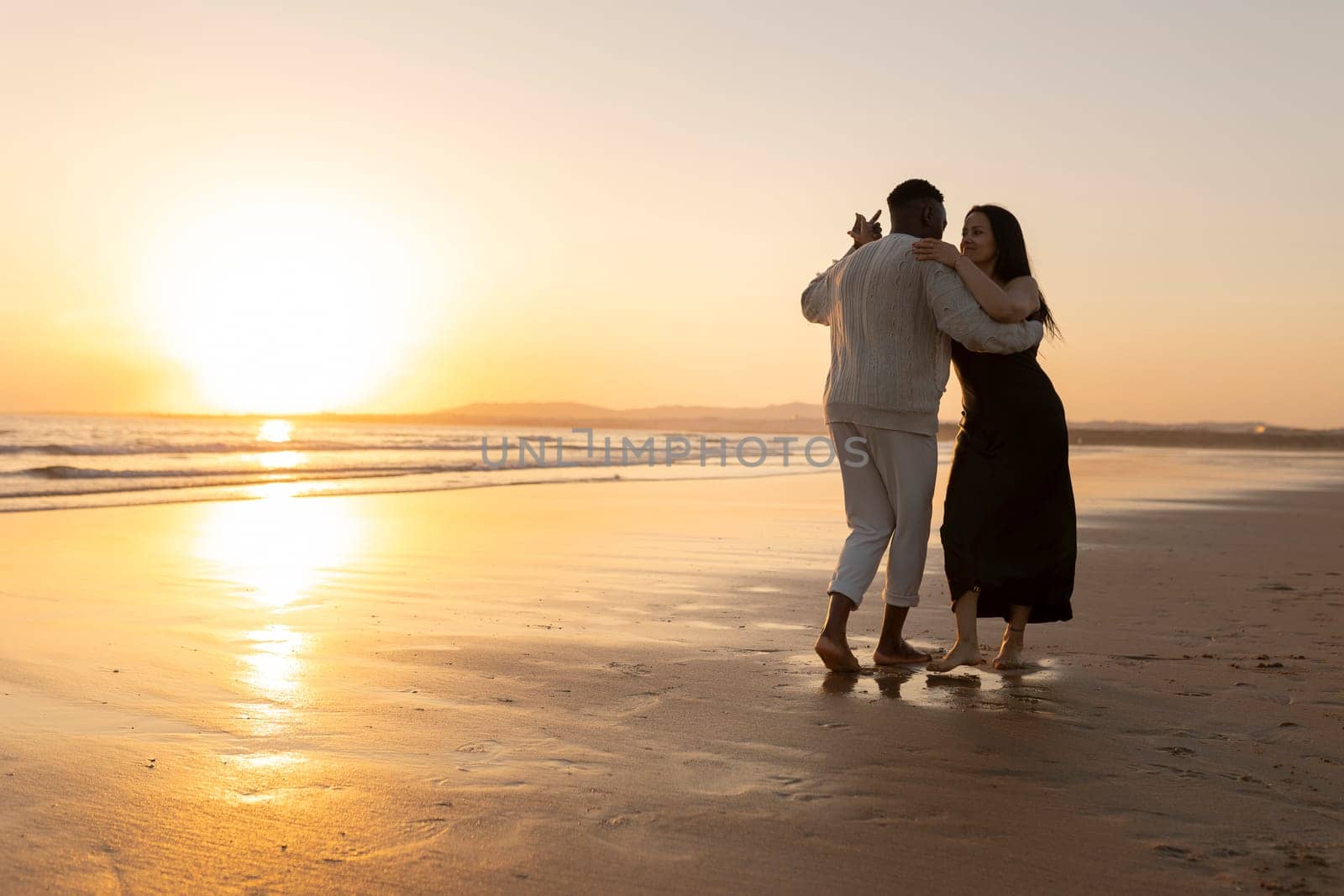 A couple dancing on the beach at sunset. The man is wearing a sweater and the woman is wearing a black dress