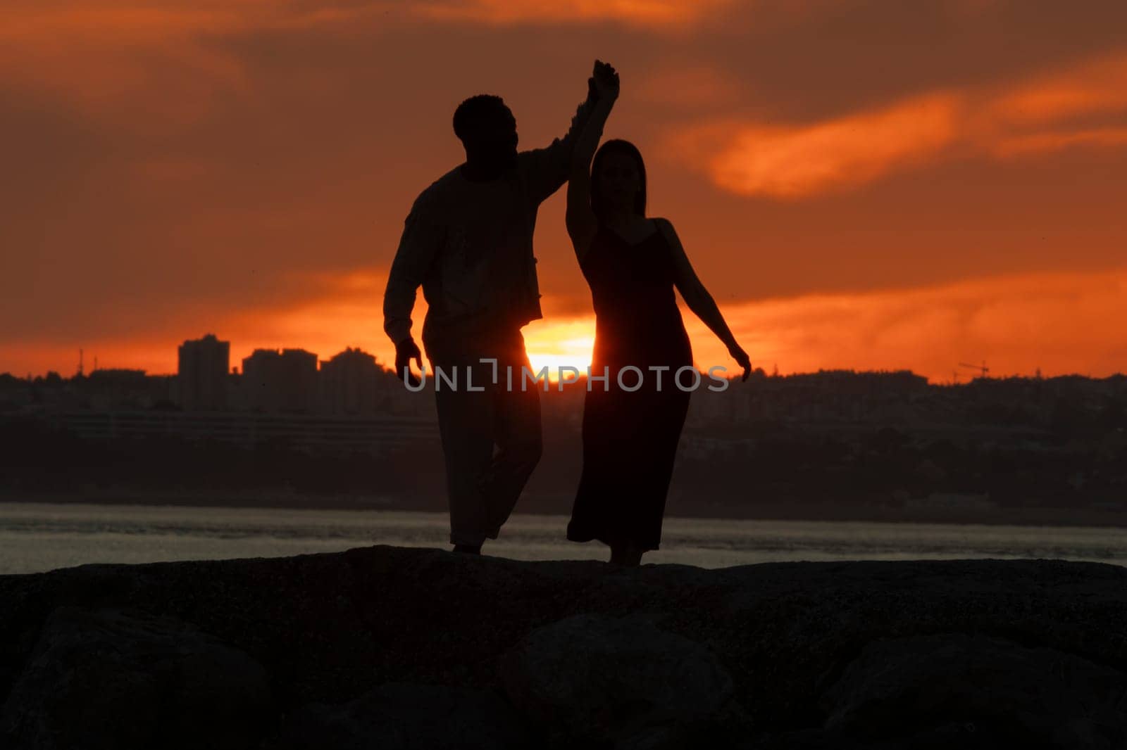 A couple dancing in the sunset. The man is holding the woman's hand. The woman is wearing a black dress