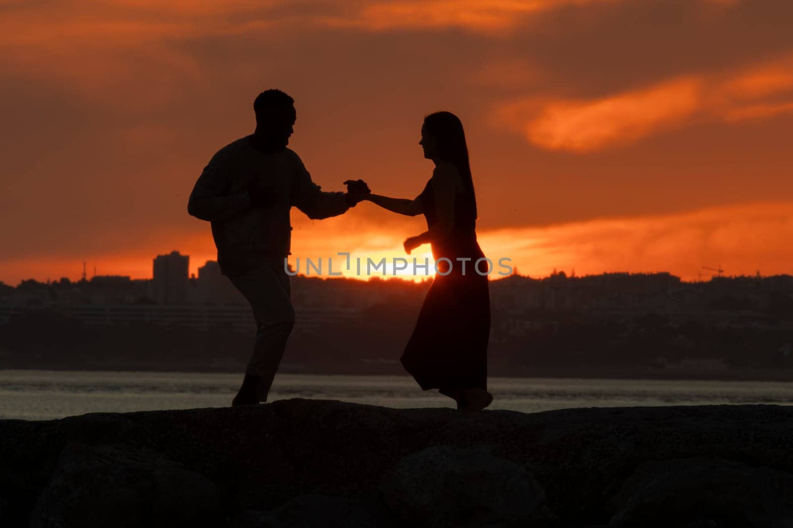 A couple dancing on a beach at sunset. The man is wearing a white shirt and the woman is wearing a black dress. The sky is orange and the water is calm