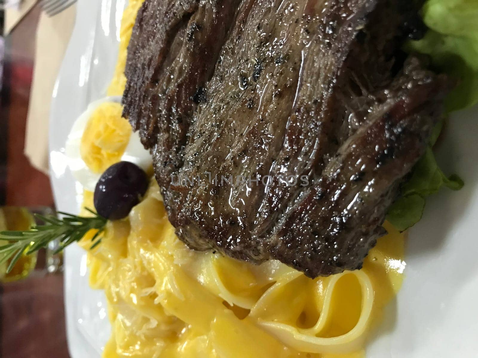 Delectable grilled beef steak served on a bed of creamy pasta, garnished with salad greens, egg, and an olive