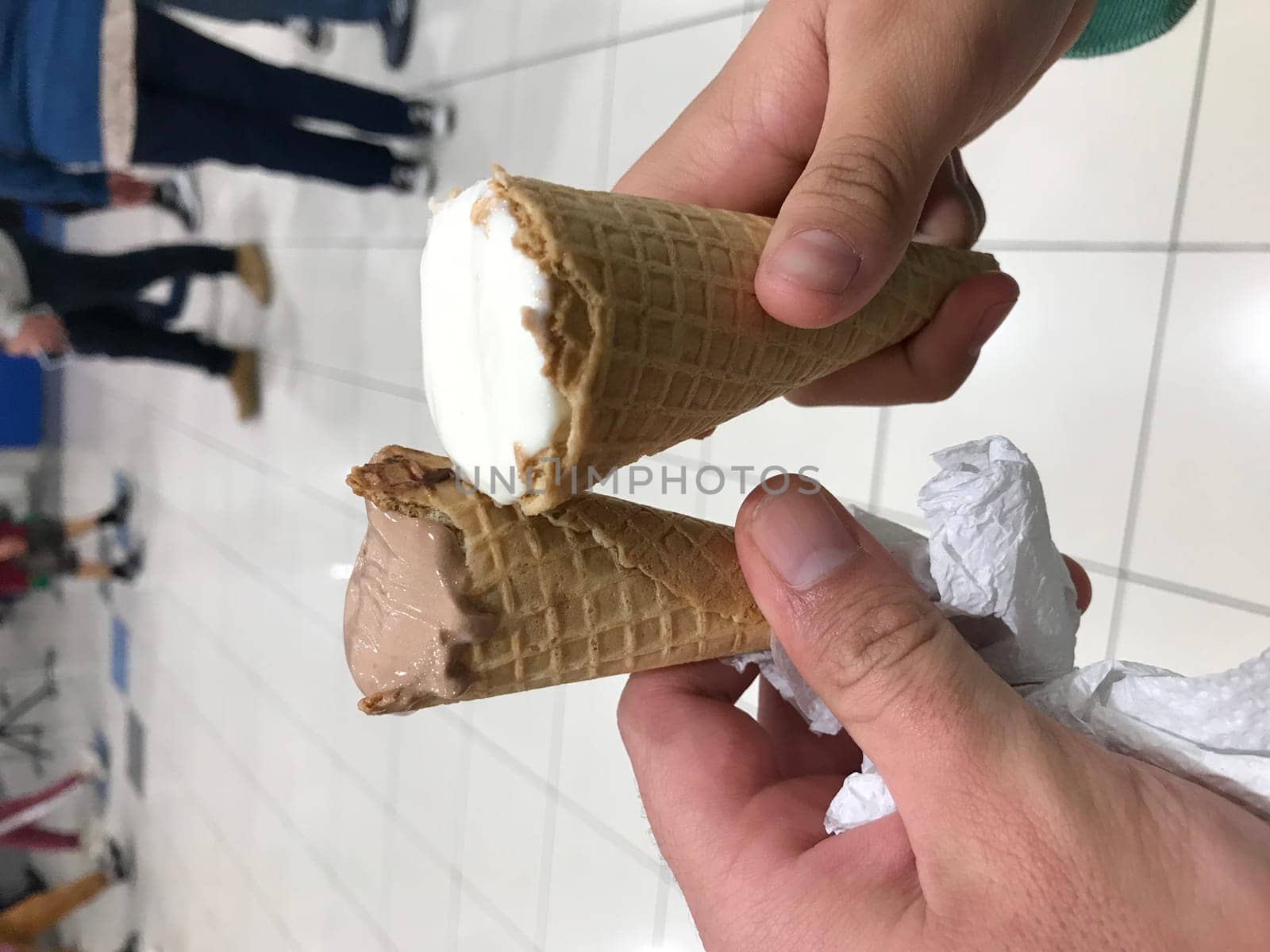 Close-up of a hand holding two ice cream cones with melting scoops, indoors with blurred people in the background