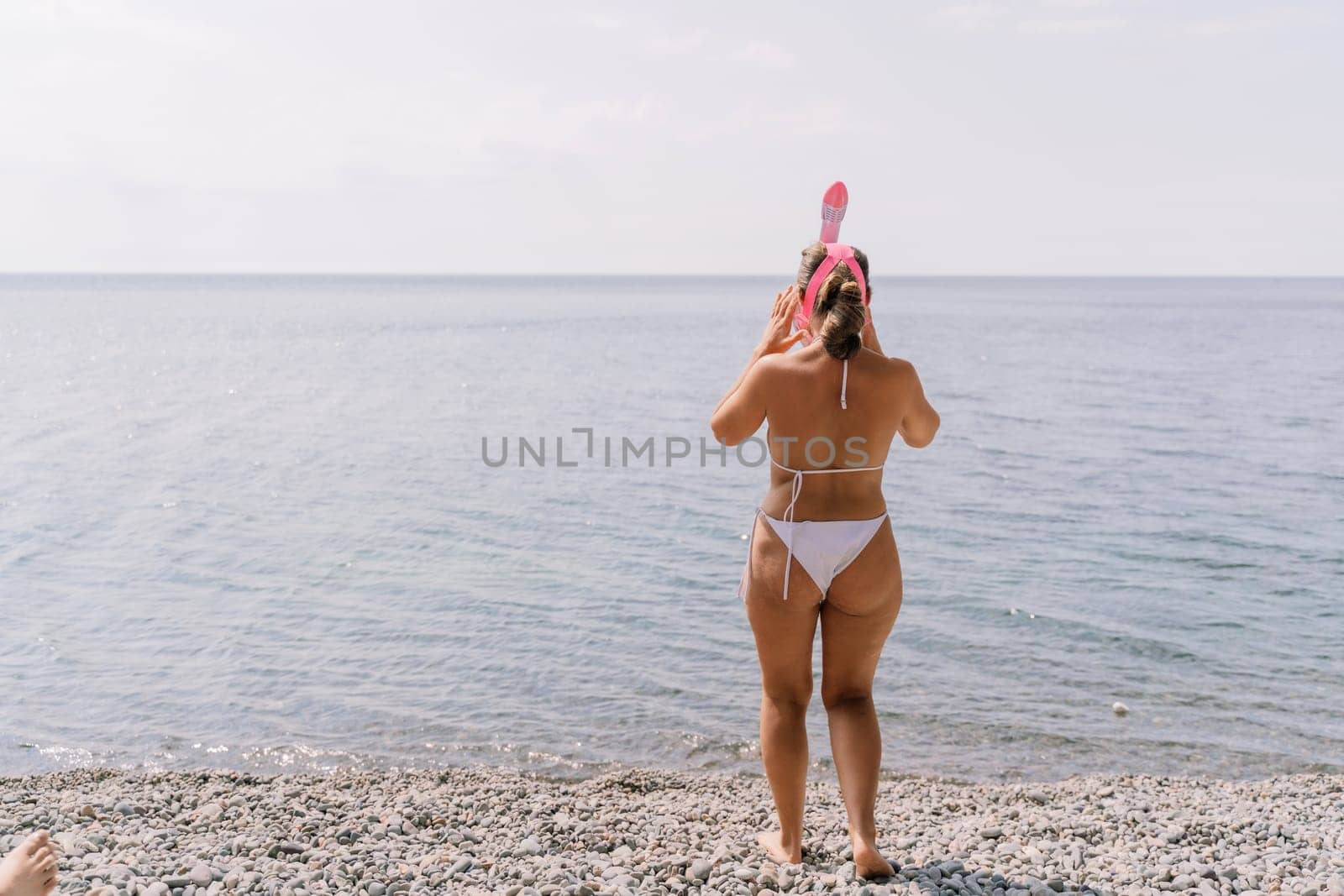 A woman in a bikini stands on a beach, looking out at the ocean. She is holding a pink snorkel and she is enjoying the view