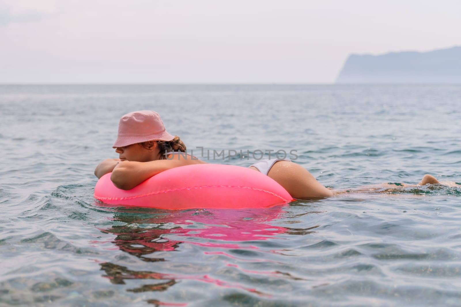 A woman is floating on a pink inflatable raft in the ocean. The water is calm and the sky is cloudy. The woman is wearing a white bikini and a pink hat