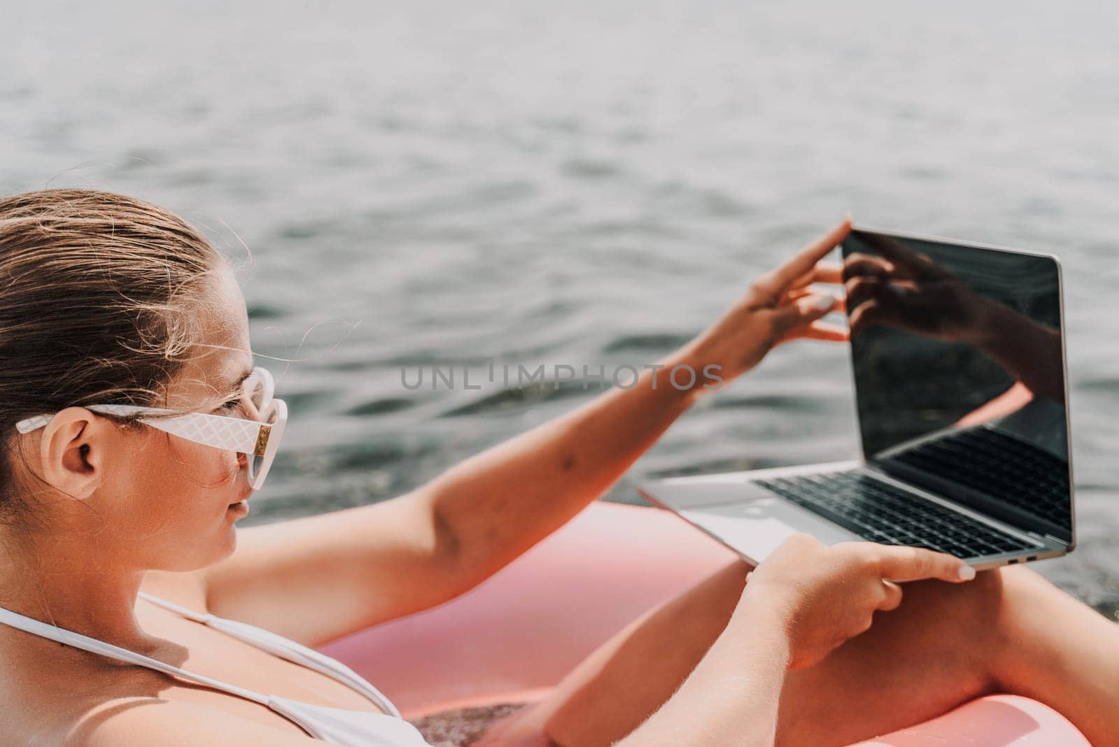 A woman is sitting in a pink inflatable raft with a laptop on her lap. She is wearing sunglasses and she is enjoying her time by the water