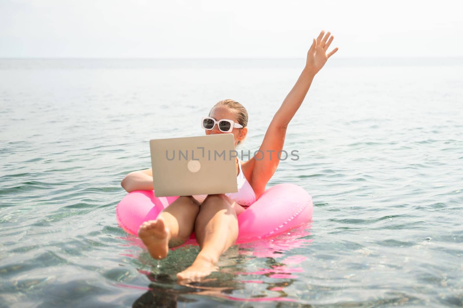 A woman is sitting on a pink inflatable raft in the ocean, holding a laptop. She is smiling and waving at the camera. Concept of relaxation and leisure