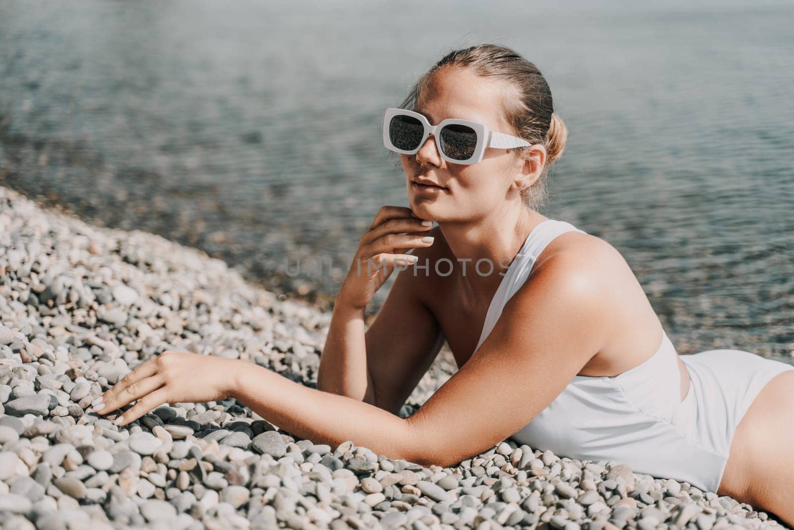 A woman is laying on the beach wearing sunglasses and a white tank top. She is looking at the water and she is enjoying the sun