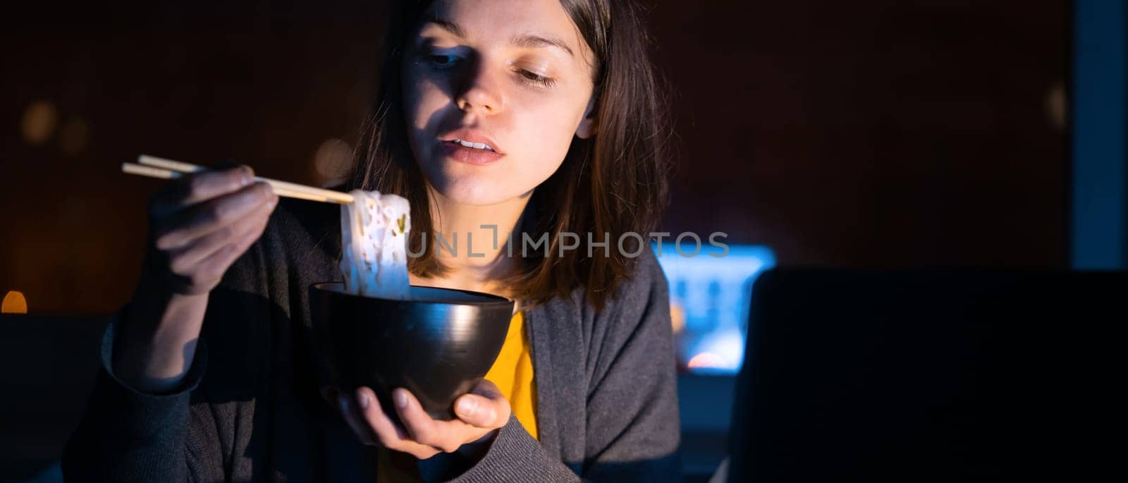 Young girl works at night at home with laptop and eats noodles, asian food with chopsticks. A woman watches a show, a movie and has dinner with ordered food from a cafe.