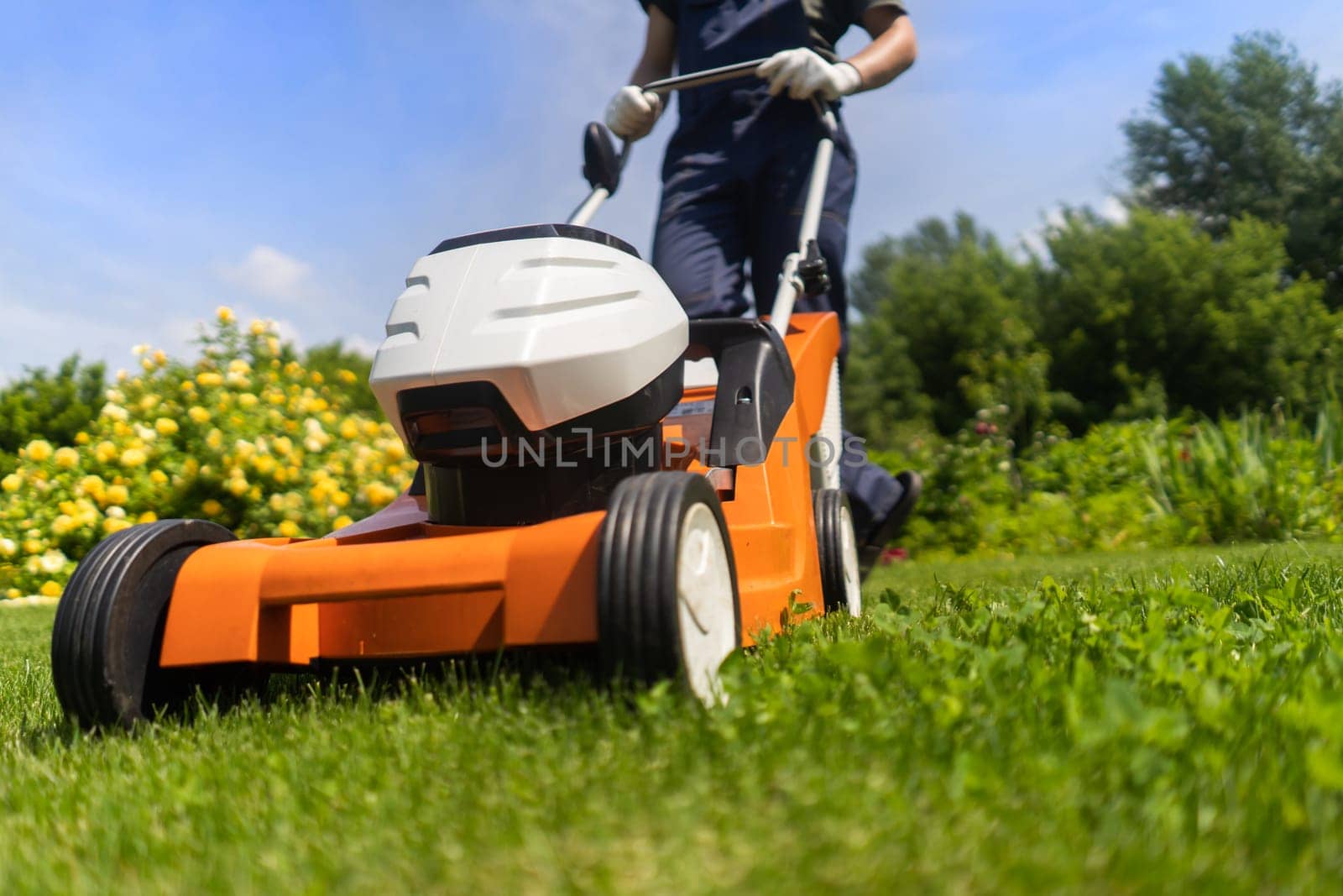 A young man in a straw hat is mowing a lawn with a lawn mower in his beautiful green floral summer garden. A professional gardener with a lawnmower cares for the grass in the backyard.