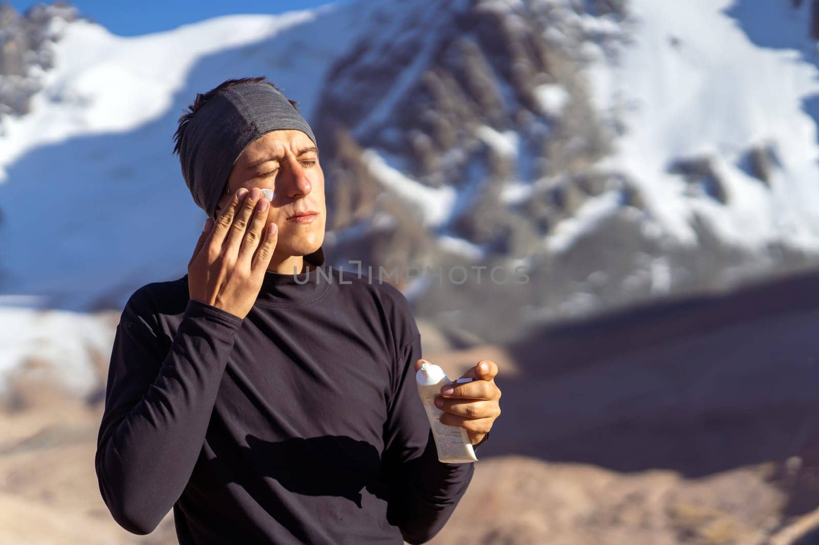 A young man applies sunscreen to his face in the highlands, in the background the peaks of the mountains are covered with snow. The traveler takes care of his skin while climbing.