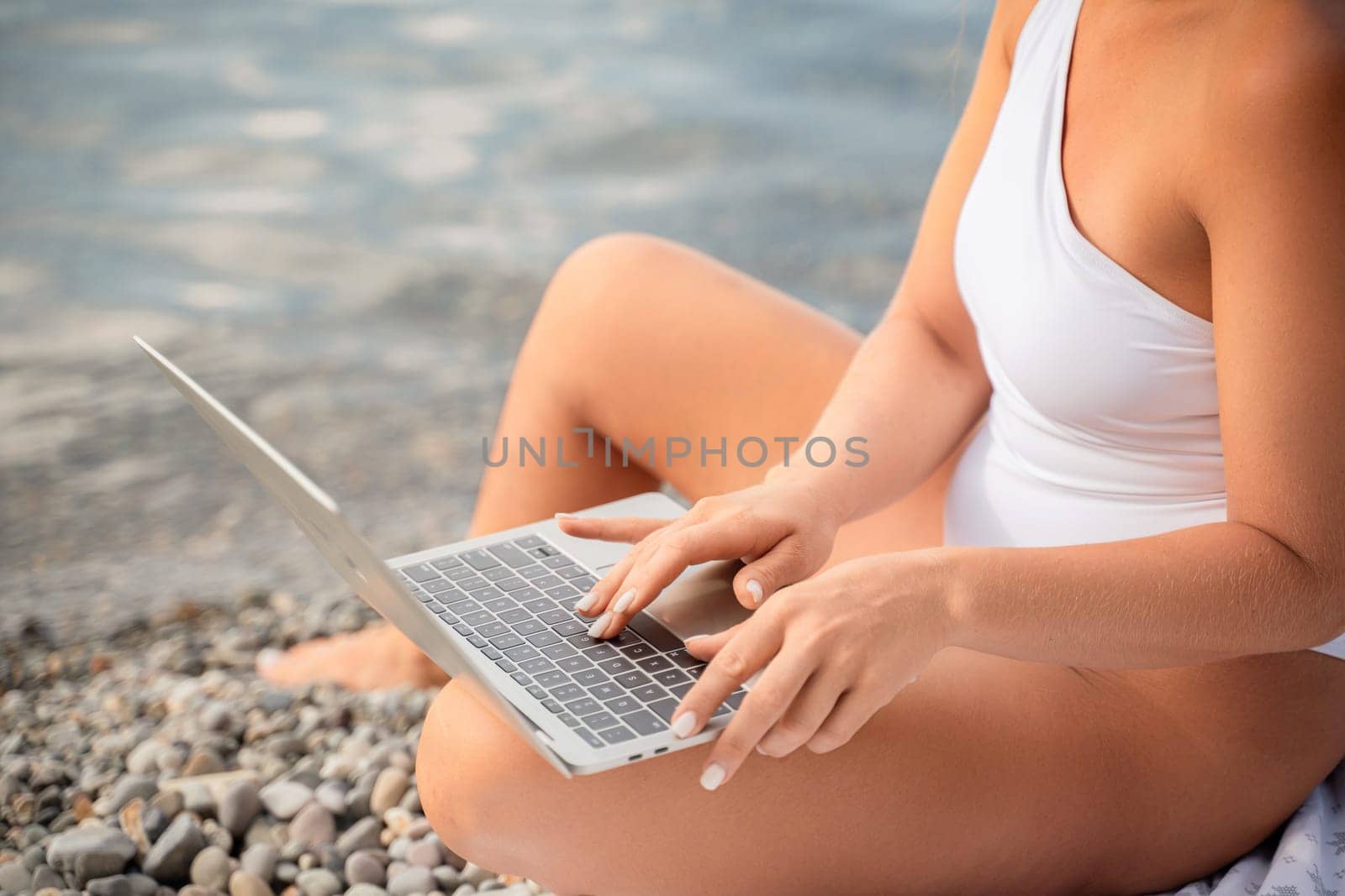 A woman is sitting on the beach with a laptop in front of her. She is typing on the keyboard and she is focused on her work