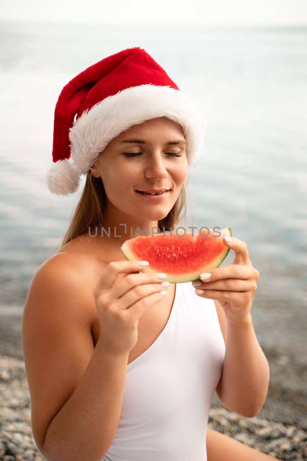 A woman is sitting on the beach and eating a watermelon. She is wearing a red santa hat and a white bikini top. Scene is lighthearted and fun