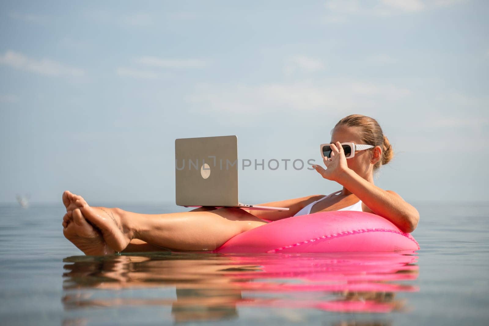 A woman is sitting on a pink inflatable raft in the water, using a laptop. Concept of relaxation and leisure, as the woman is enjoying her time in the water while working on her laptop